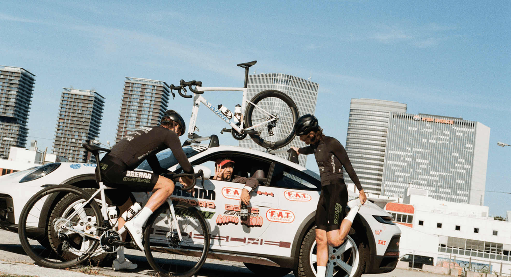 Two cyclists in black aerodynamic racing kits with 'Dream' branding adjust their white road bikes beside a branded white car.