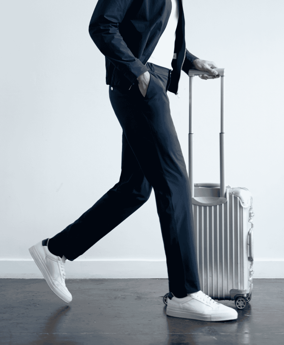 Full-body shot of a man in slim-fit navy trousers and white sneakers walking with luggage.