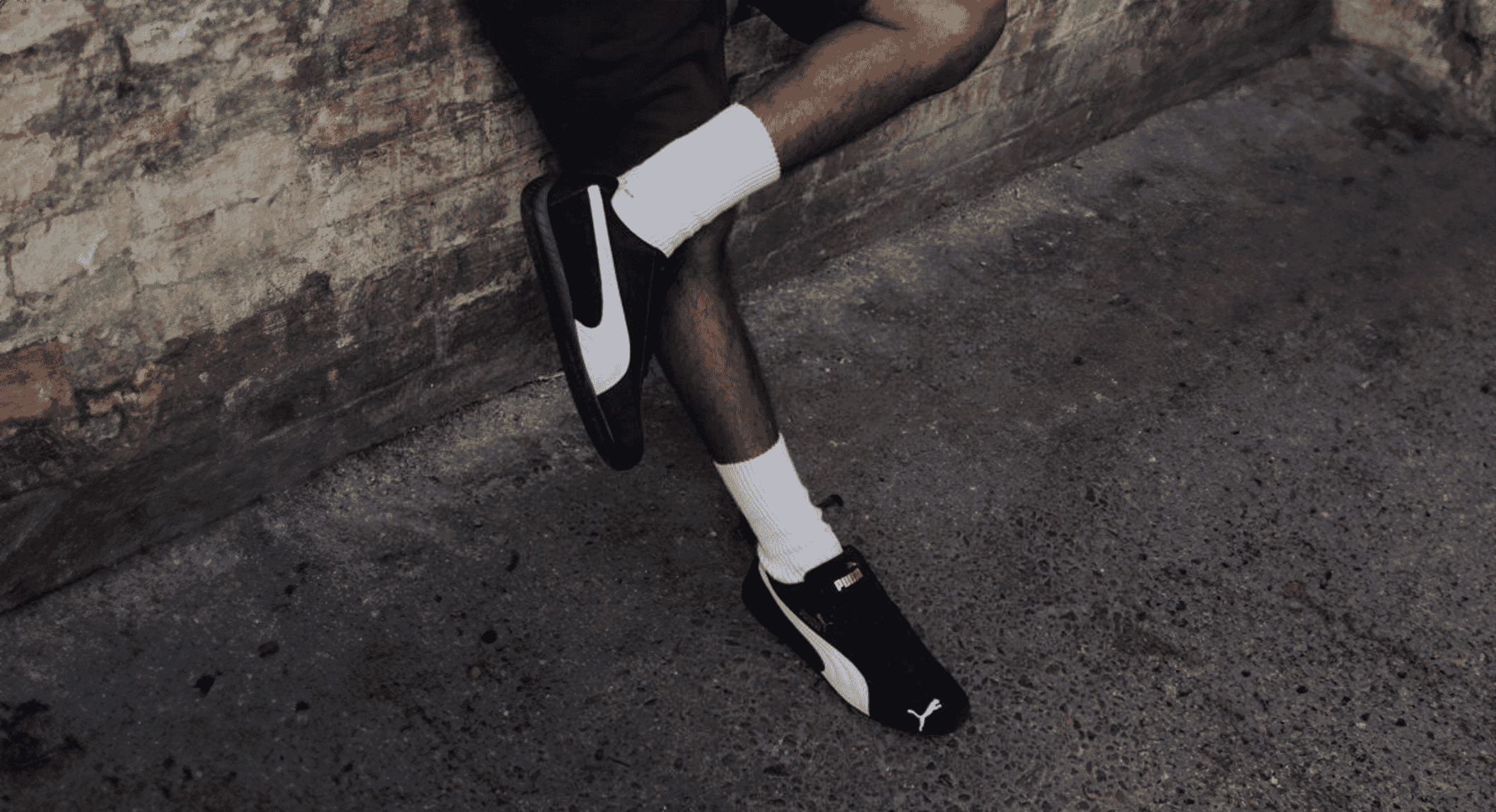 Streetwear shot of a person in black slim Puma sneakers against an urban brick wall.