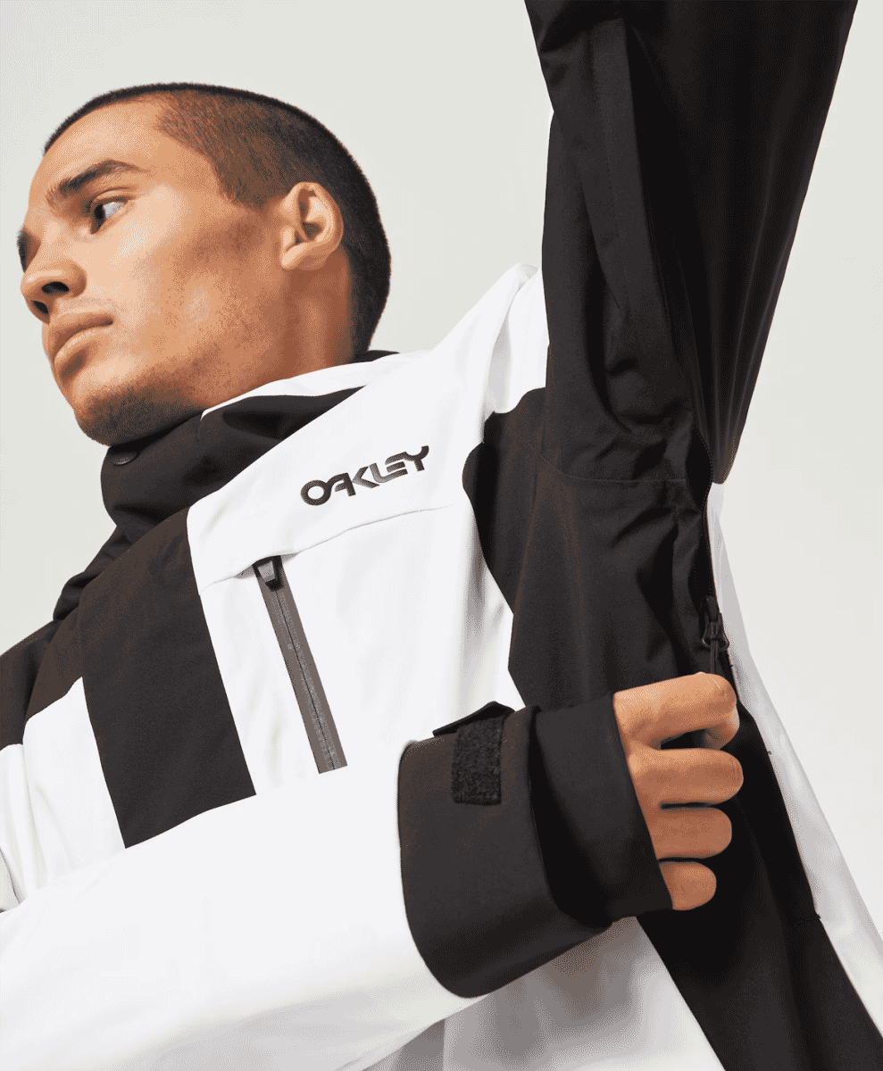 A studio shot of a model adjusting the sleeve of a black and white Oakley technical ski jacket.