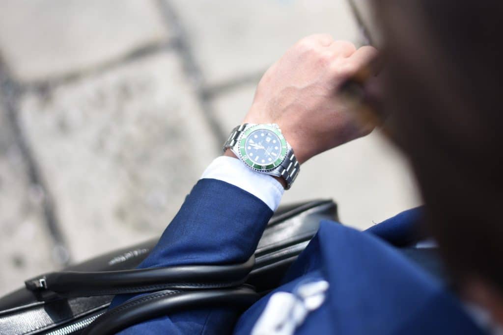 A man in a navy blue suit checking a green-bezel Rolex watch in a city environment.