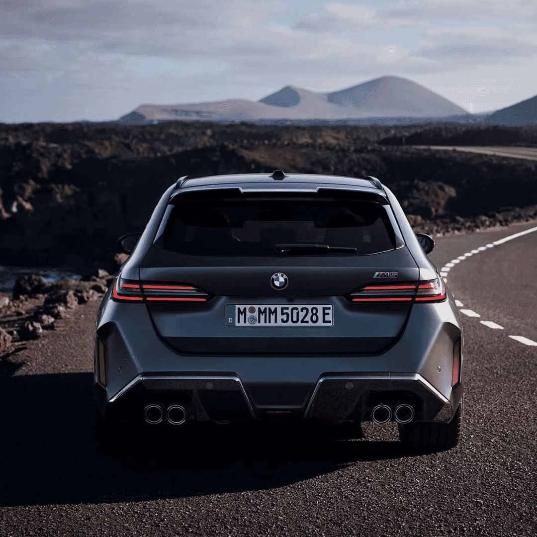 Rear view of a metallic gray BMW M5 Touring on a winding mountain road, showing the dual exhaust outlets and wide rear arches.