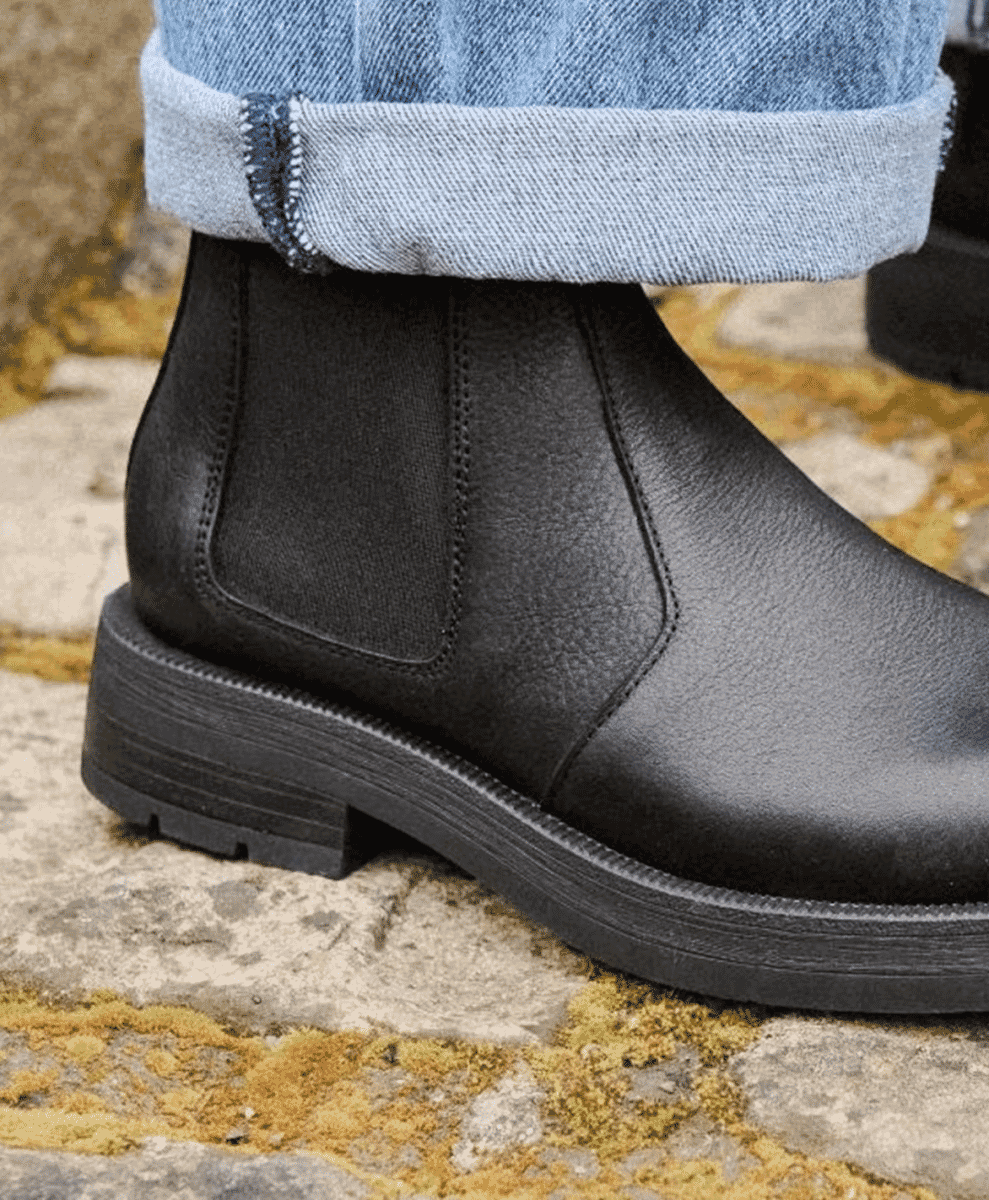 Close-up of black leather Chelsea boots standing on a wet, mossy stone surface.