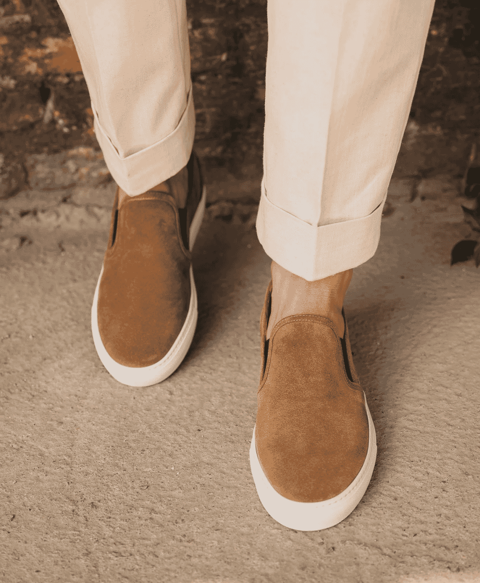 Close-up of tan suede slip-on sneakers paired with cuffed beige trousers.