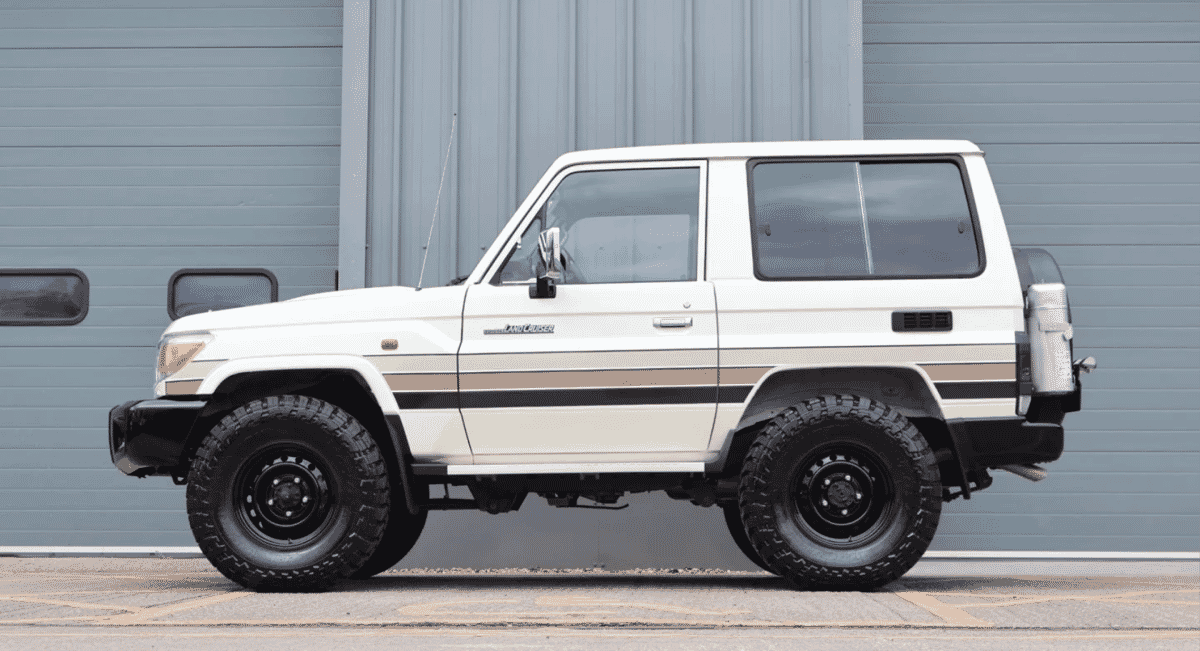 A white vintage Toyota Land Cruiser with beige stripes parked in front of a gray industrial garage.