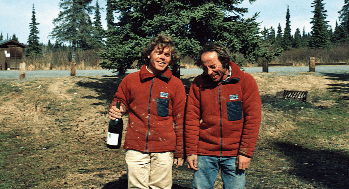 Two men in matching red fleece jackets smiling in a rustic outdoor setting.
