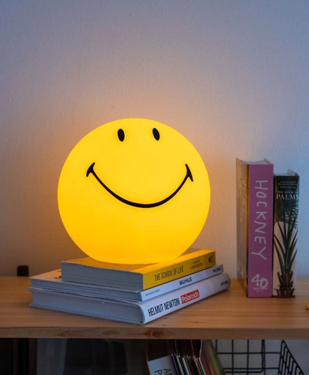 Glowing yellow smiley face lamp resting on a stack of books on a shelf.