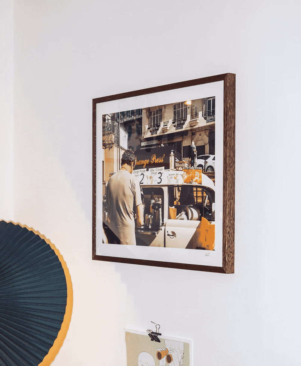 Framed photo of a man at an orange juice cart on a sunlit European street.