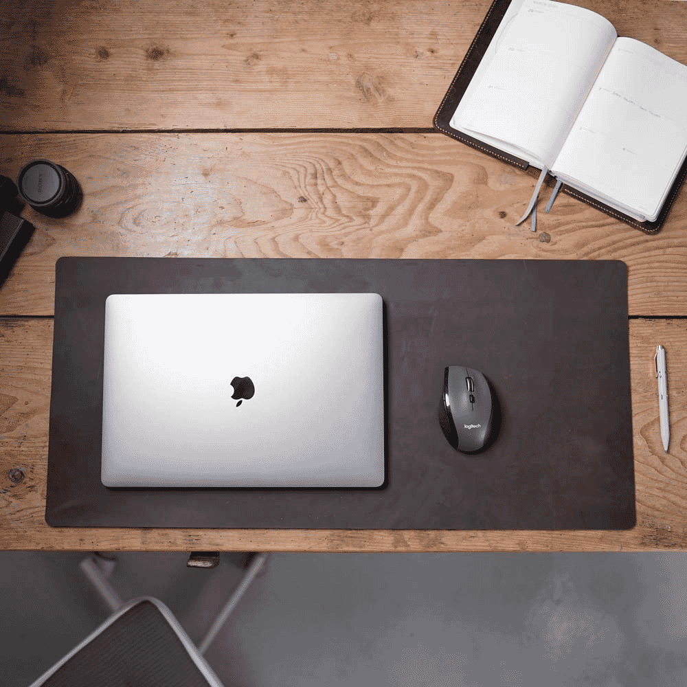 Top-down view of a minimalist wooden desk featuring a laptop, notebook, and a sleek dark desk mat.