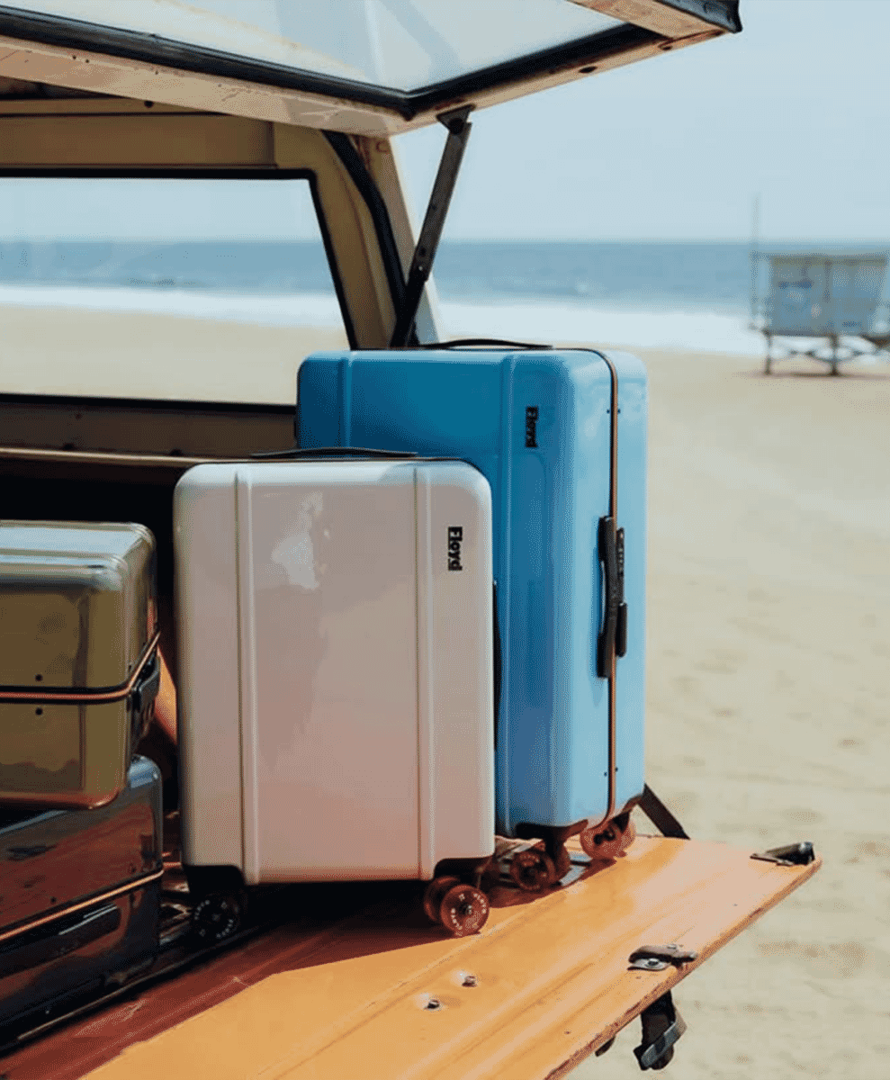 Three stacked Floyd brand suitcases in white, blue, and metallic in a vintage van trunk