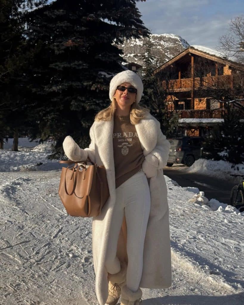 Woman in a white shearling coat and Prada sweatshirt in an alpine village.