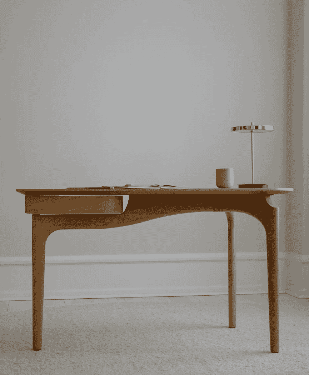 Minimalist light oak desk with sculpted legs and a single drawer against a neutral wall.