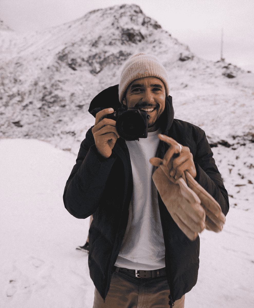 A man in winter gear holding a camera on a snowy mountain landscape.