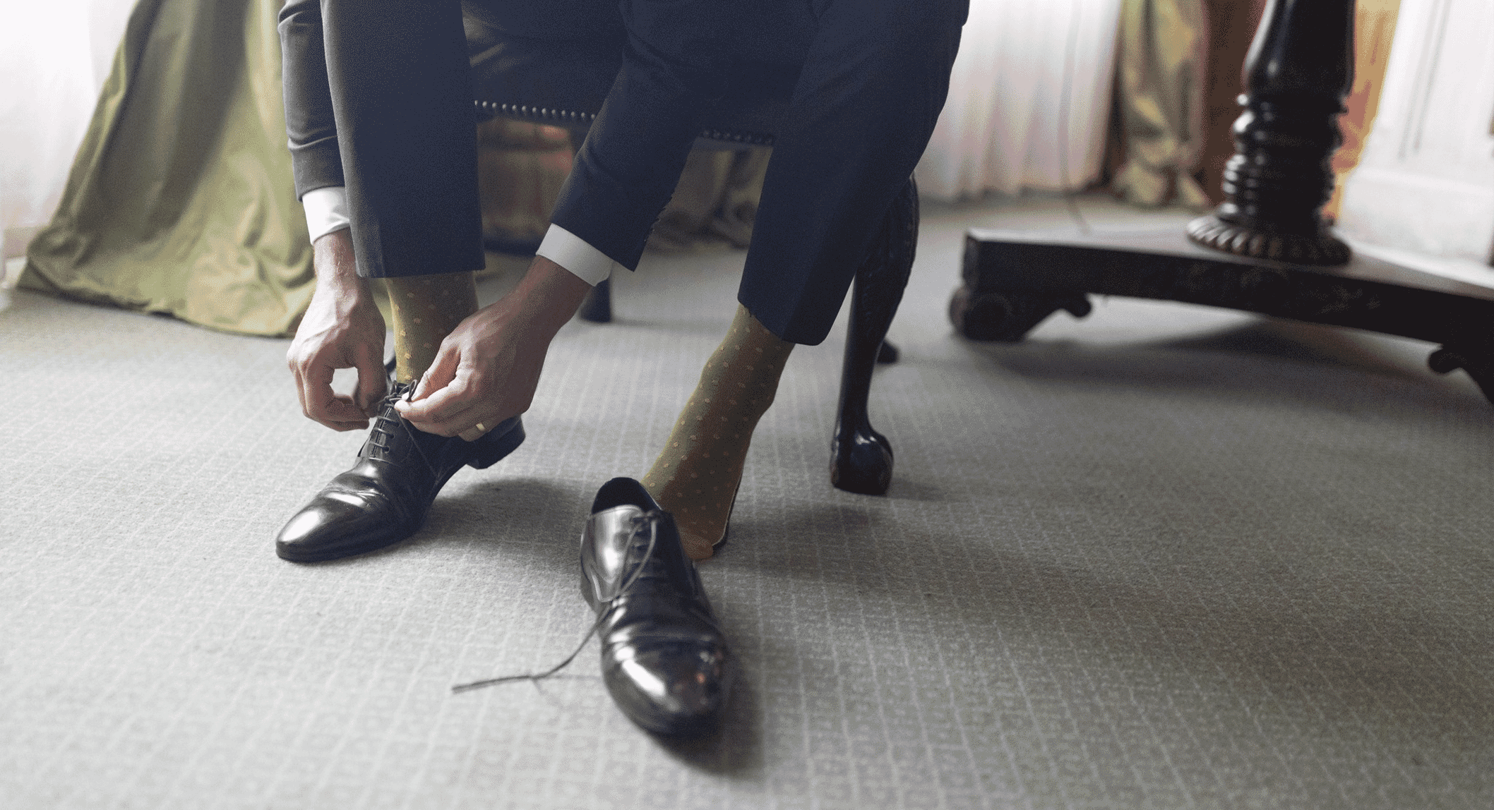 A man in a dark suit and patterned socks tying his black dress shoes in an elegant room.