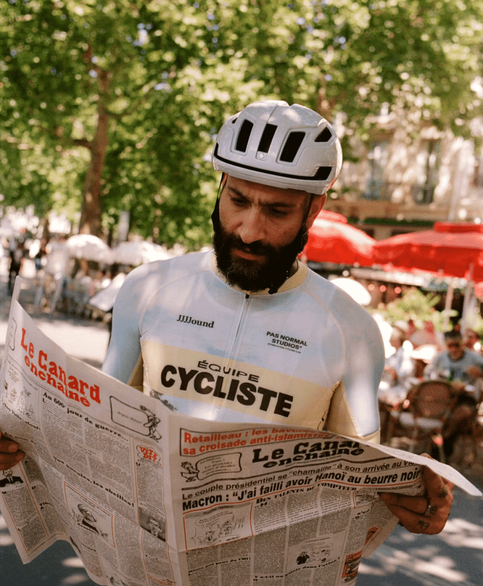 A man in a cycling jersey and white helmet reading a newspaper at an outdoor café.