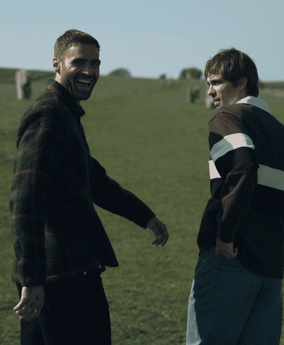 Two men outdoors, one wearing a striped rugby-style jacket and the other in a dark plaid coat.