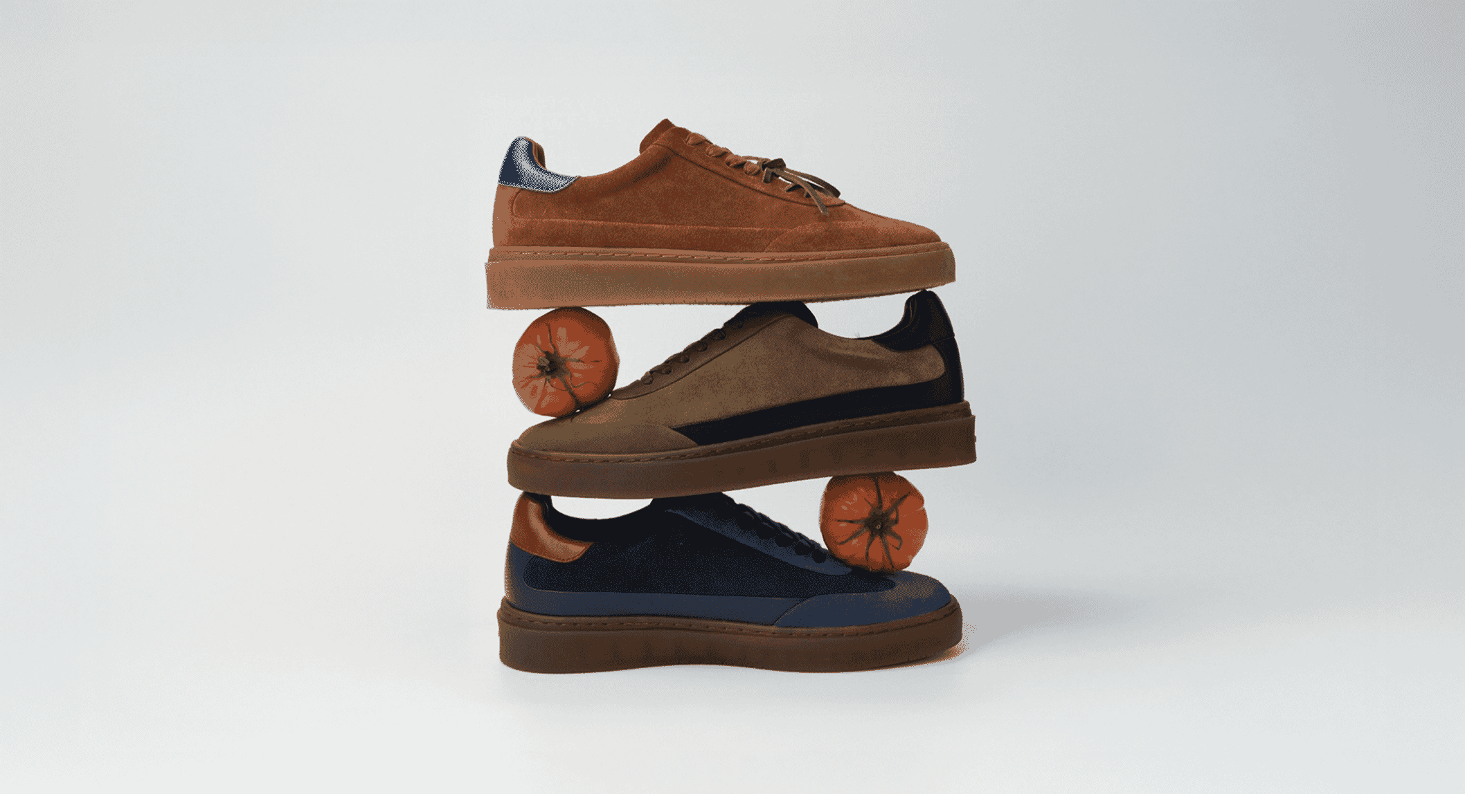 Three stacked suede sneakers in earthy tones balanced on small orange tomatoes against a white background.