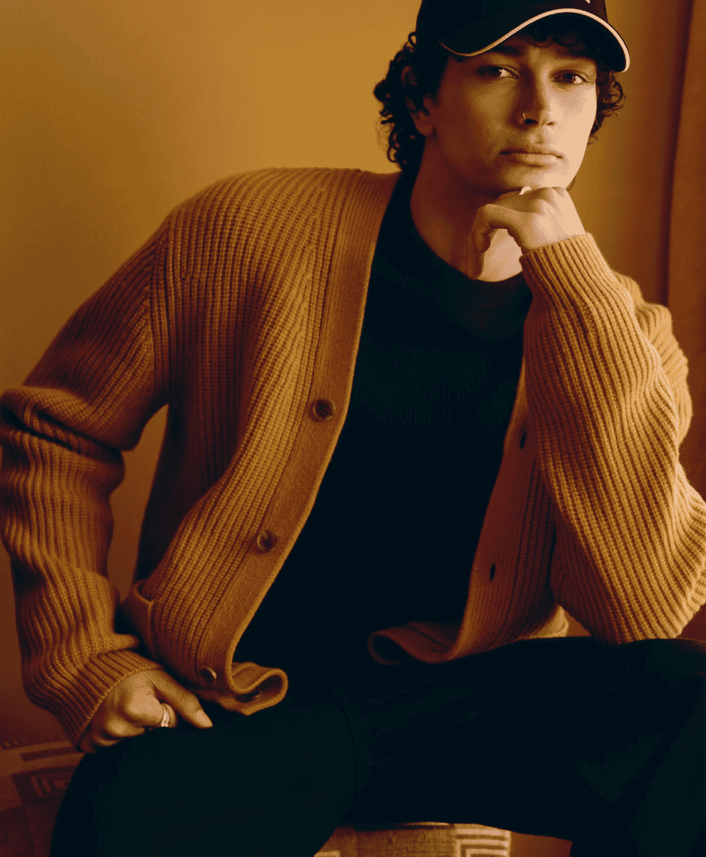 A man with curly hair wearing a ribbed brown cardigan in a thoughtful pose under warm lighting.
