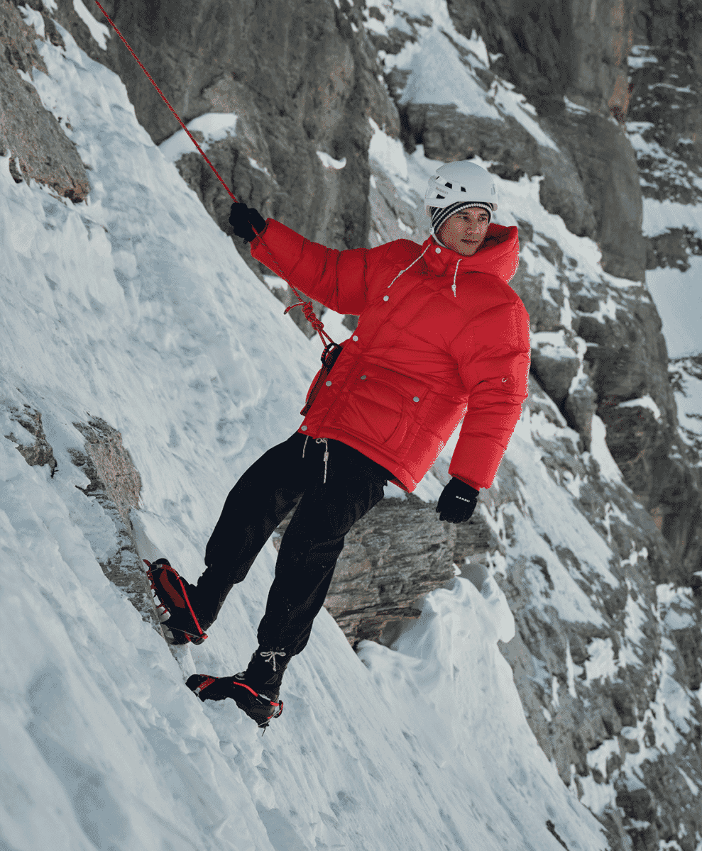 Climber in a bright red puffer jacket and white helmet scaling a snowy rock face.