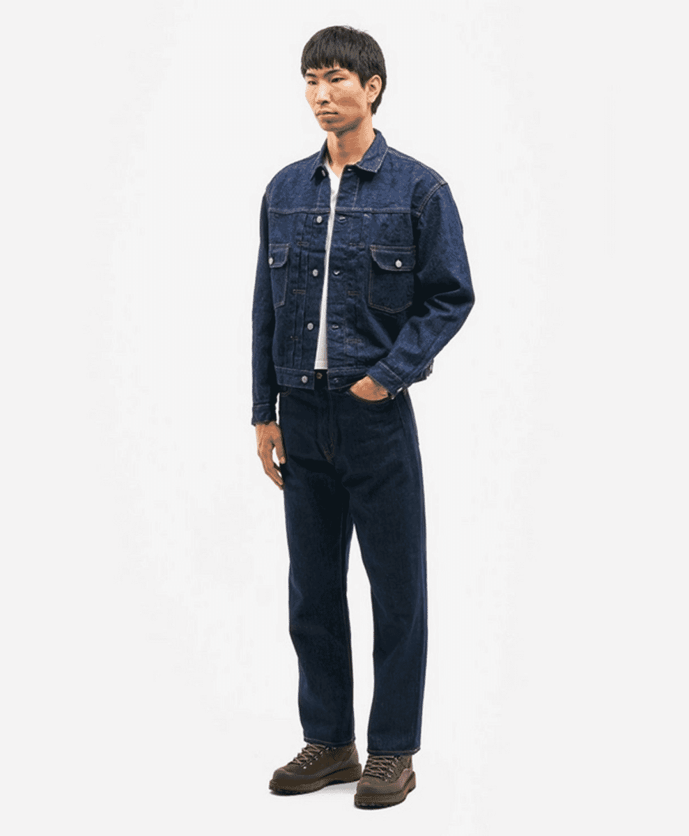 Full-body shot of a man in a dark blue denim trucker jacket and matching denim pants with brown boots.