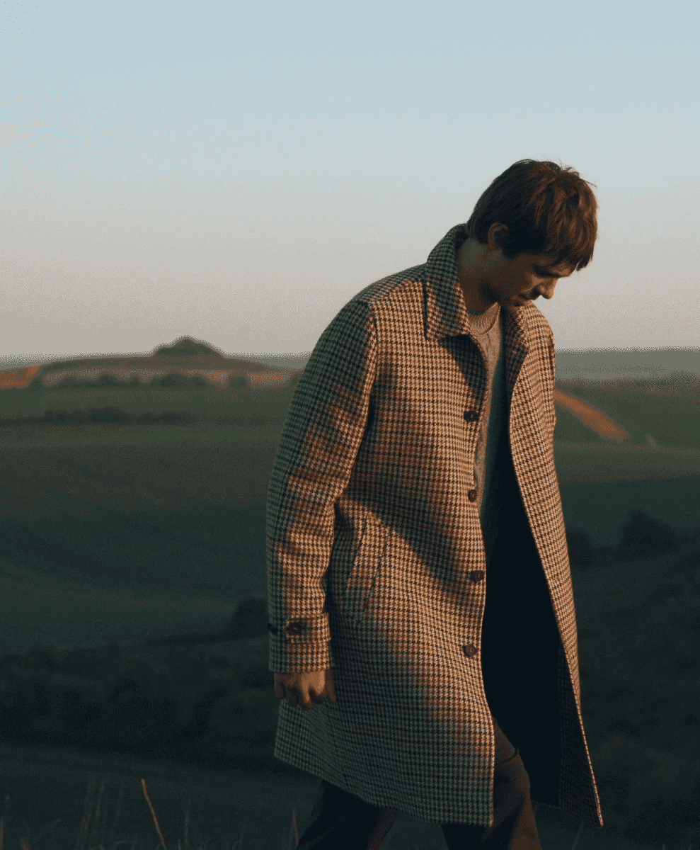 A man in a houndstooth-patterned wool coat walking across a rural hillside at golden hour.