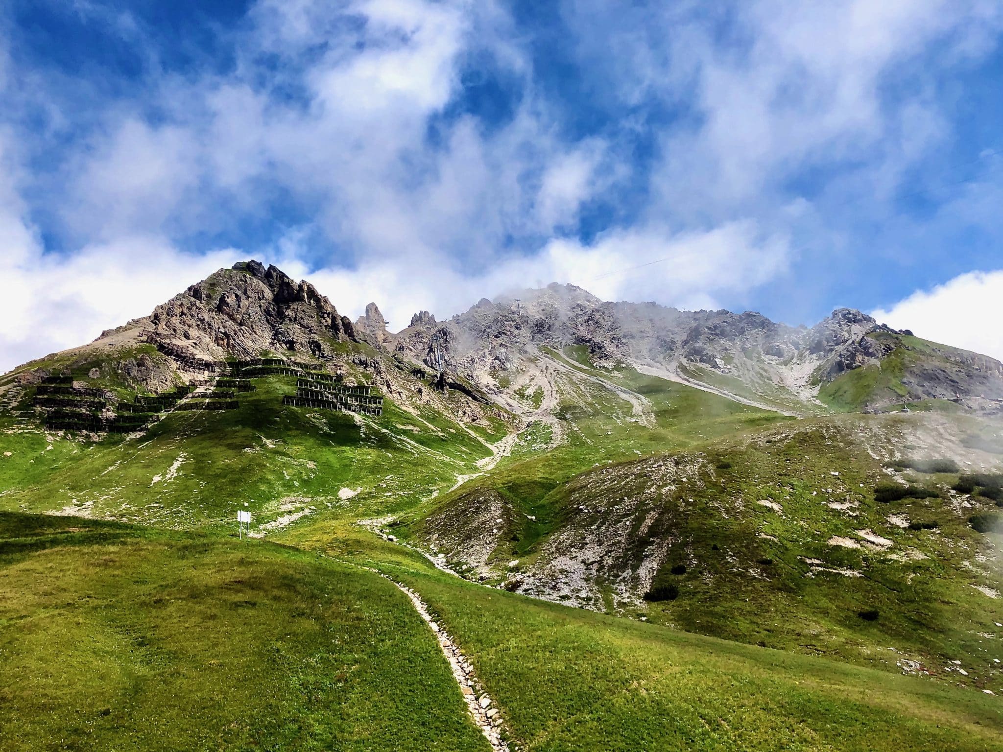 Lush green Alpine slopes leading up to jagged rocky peaks under a cloudy sky.