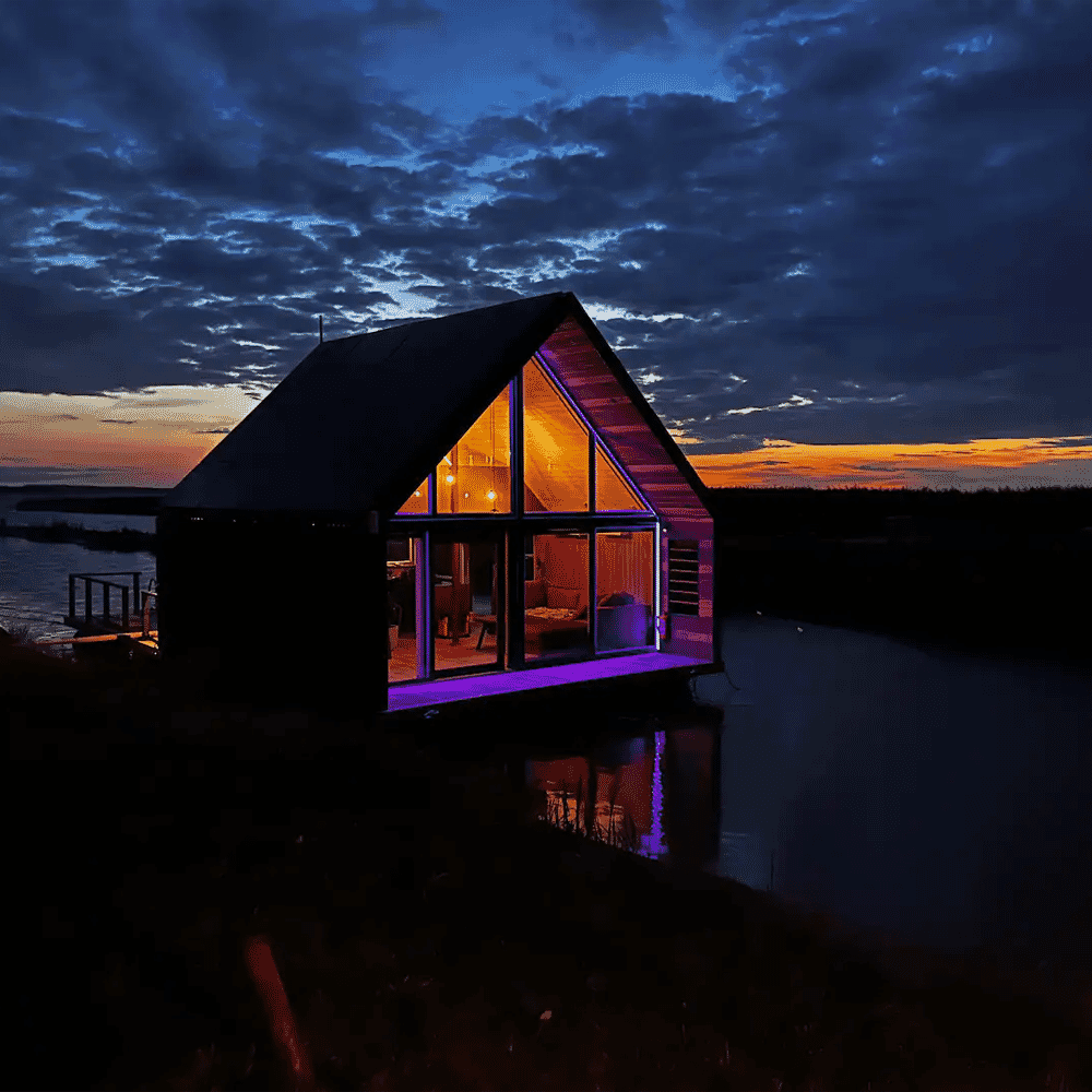 Cozy modern cabin glowing with warm interior lights by a lake at dusk under a twilight sky.