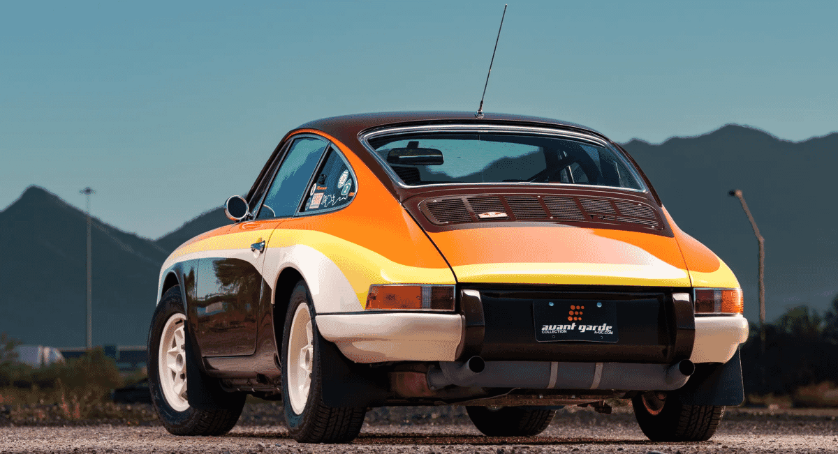 A vintage Porsche rally car with orange and yellow racing livery parked outdoors against a mountain backdrop.