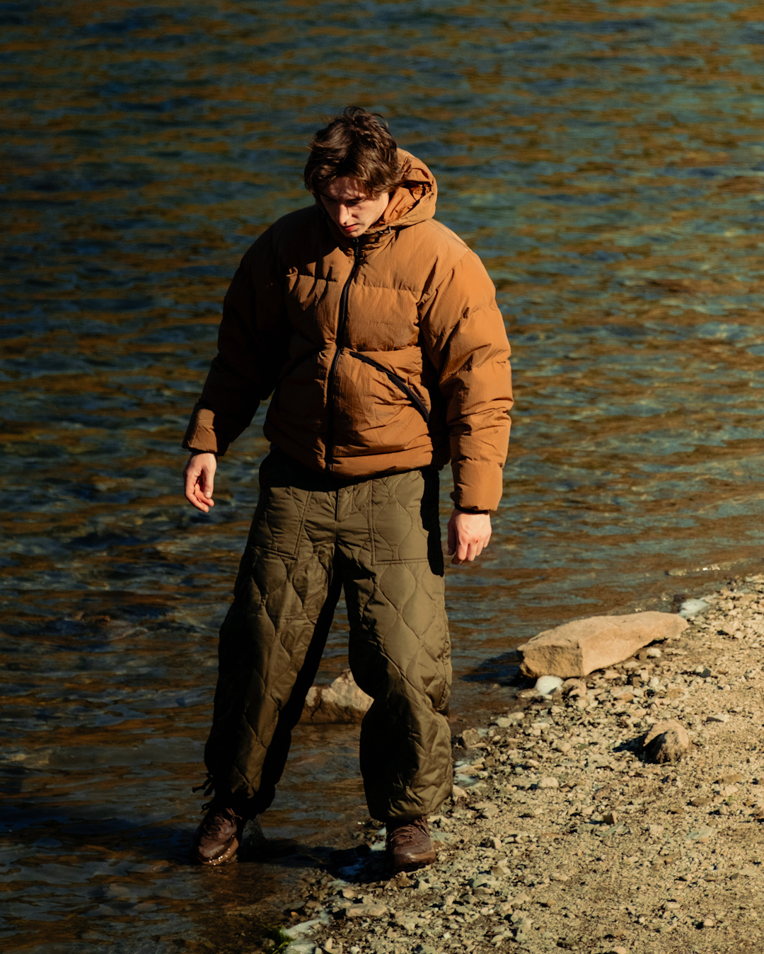 A man in a bulky brown puffer jacket walking along a rocky shoreline reflected in golden light.