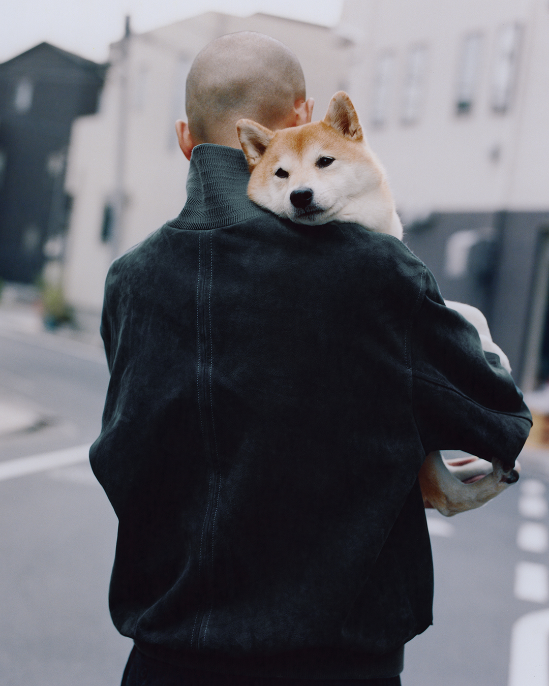Rear view of a man in a dark suede utility jacket carrying a Shiba Inu on an urban street.