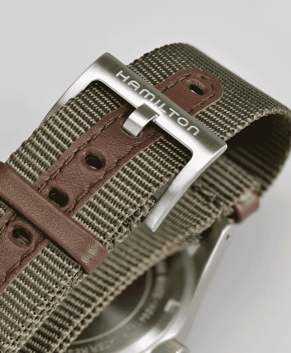 Macro shot of a Hamilton watch strap showing olive green woven fabric and a metal buckle.
