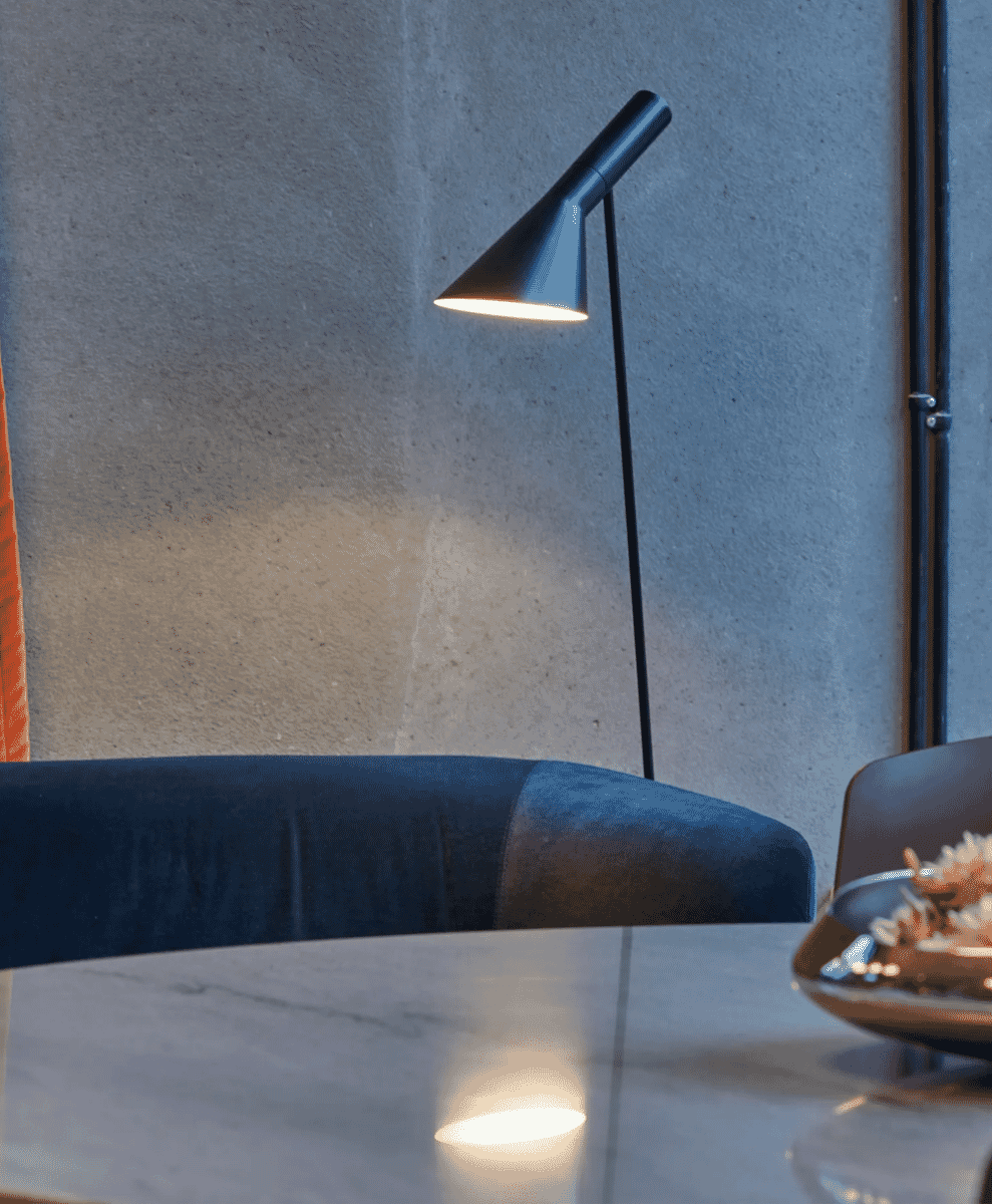 A sleek black floor lamp illuminating a marble side table next to a dark blue upholstered chair and concrete wall.