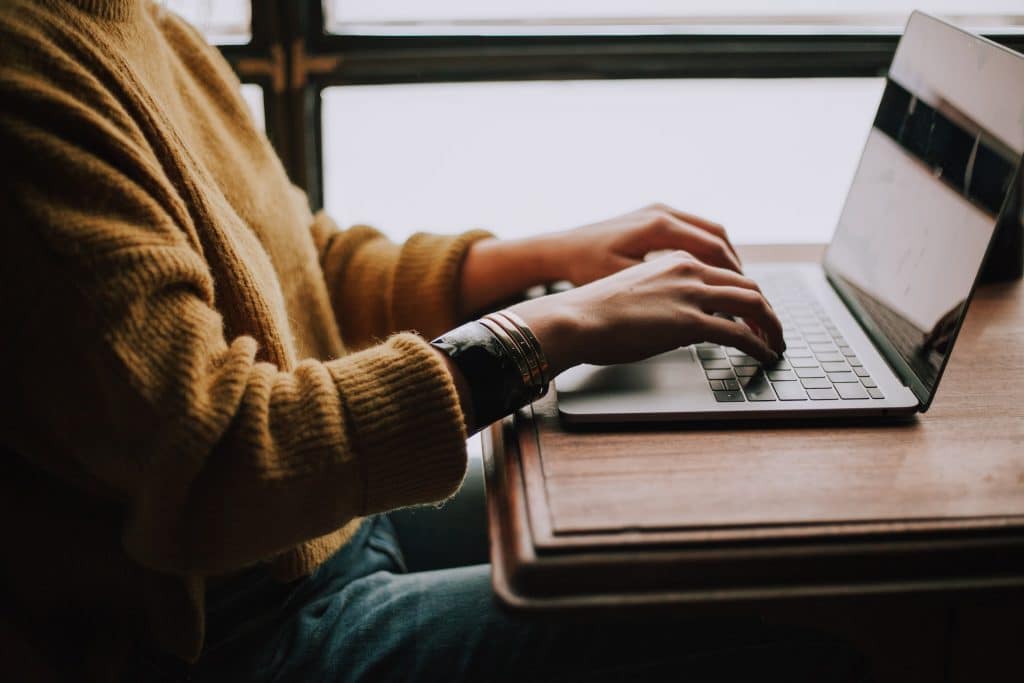 A close-up shot of a person in a mustard-yellow ribbed sweater using a laptop at a wooden desk.