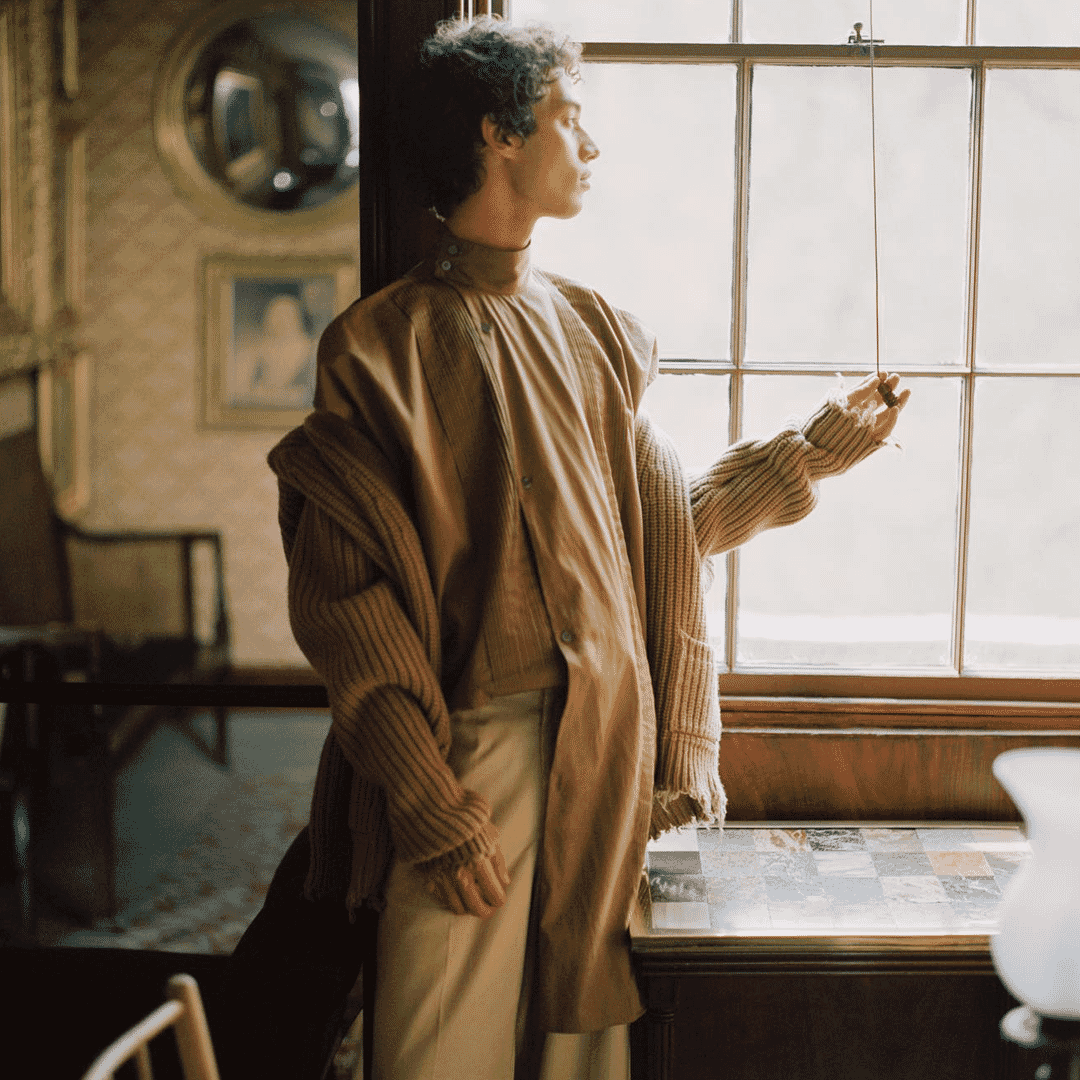 Man in a brown corduroy coat over a ribbed knit sweater standing by a window.