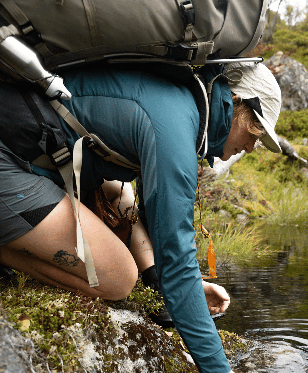 A hiker in teal gear kneeling by a stream to drink water in a rugged landscape.
