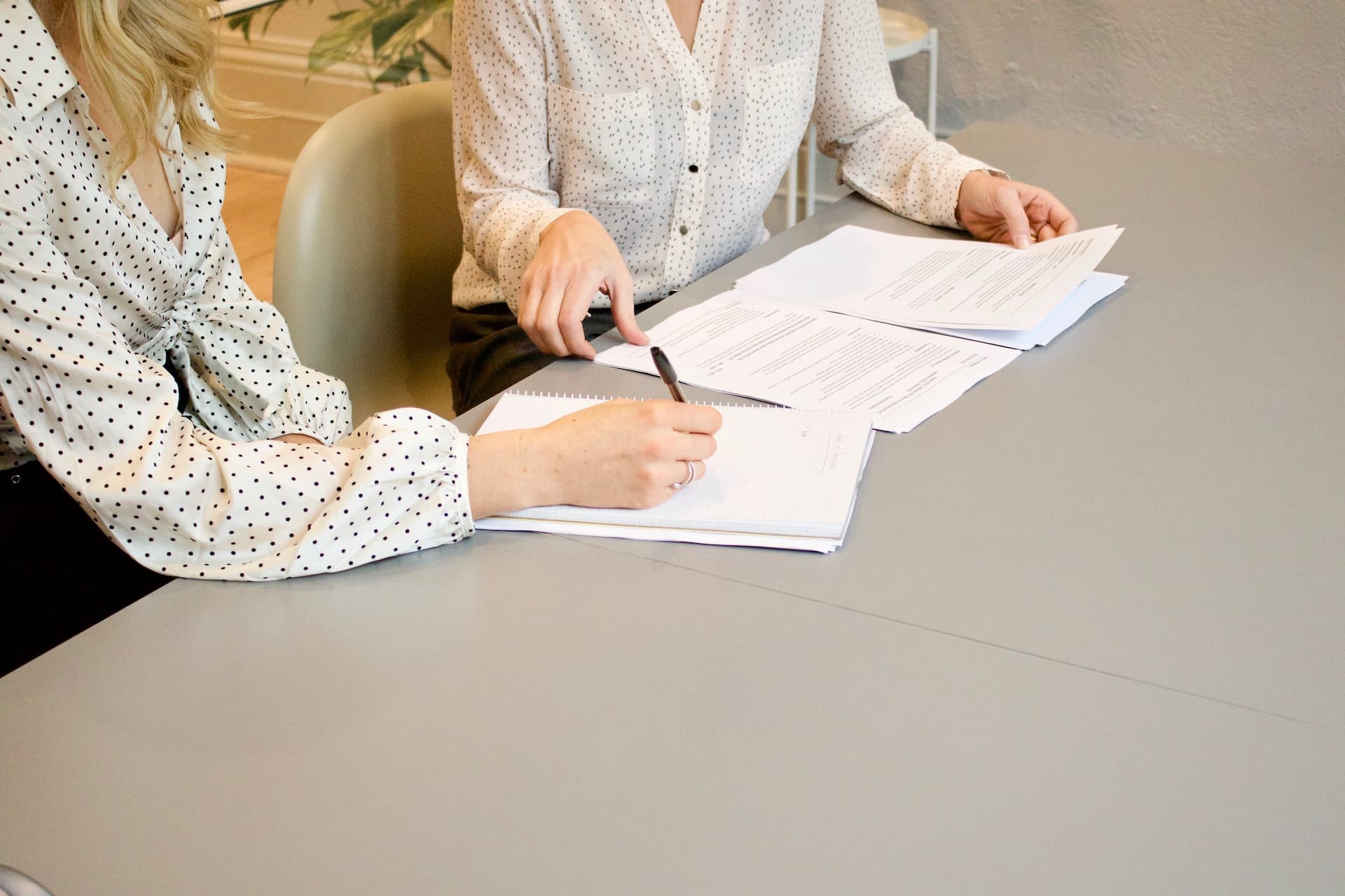 Two women in white polka-dot blouses reviewing documents and taking notes in a professional office setting.
