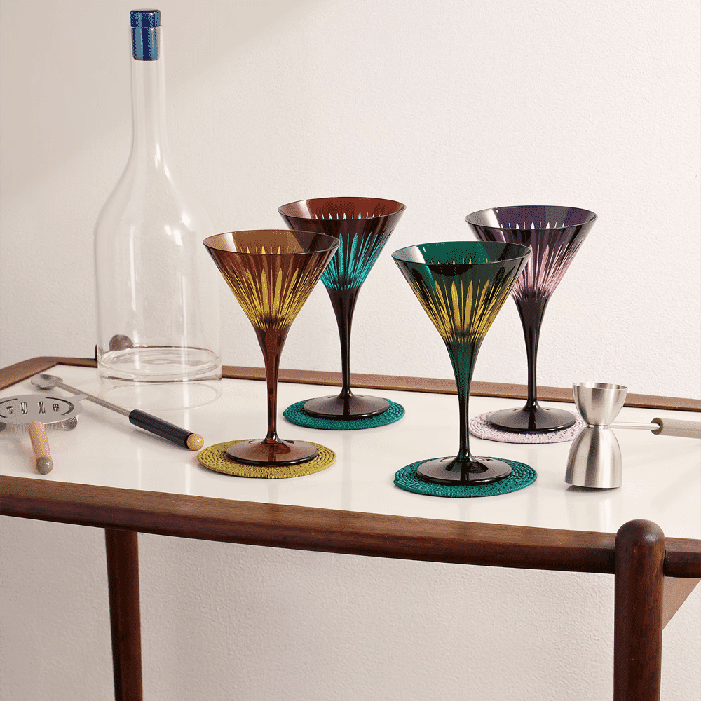 A wooden bar cart featuring a variety of colorful, patterned martini glasses and a decanter.