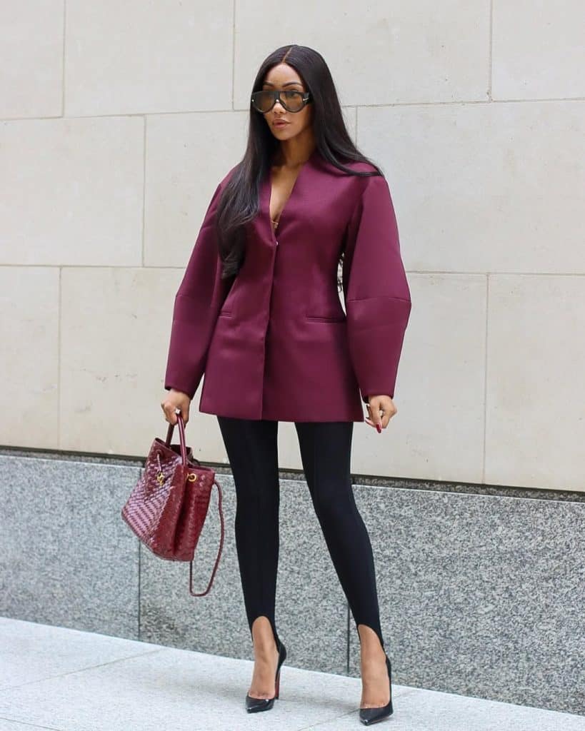 A woman wearing an oversized burgundy blazer and black leggings posing against a modern stone wall.