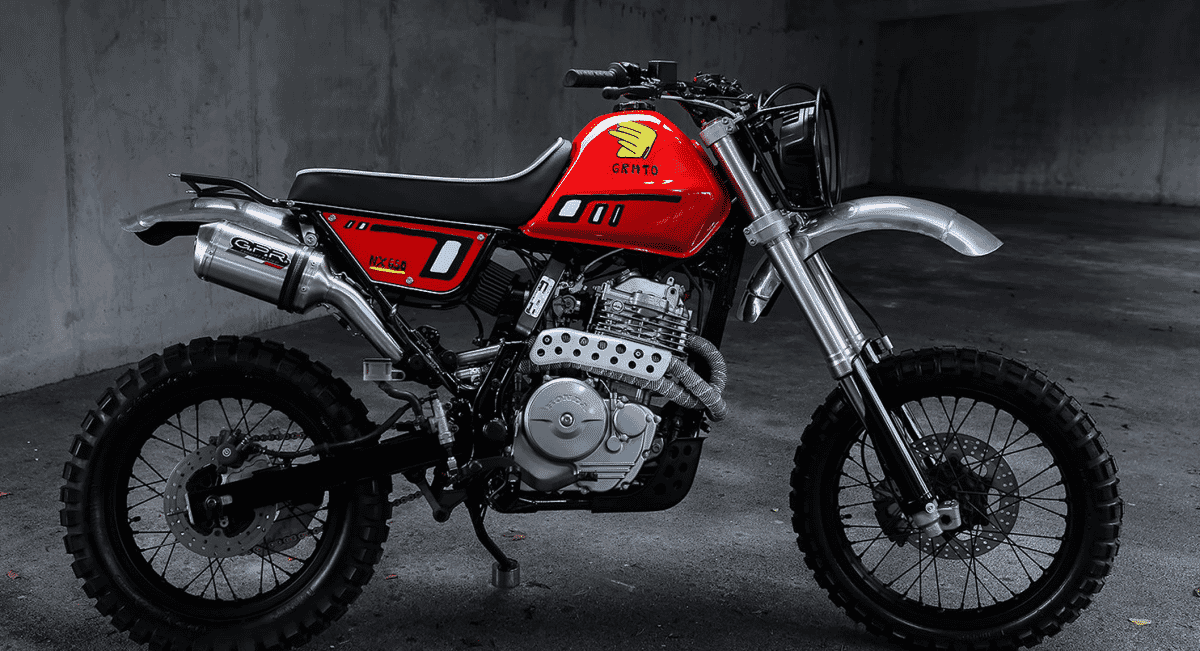 Wide-angle shot of a custom red and silver Honda motorcycle with rugged tires in a monochrome industrial setting.