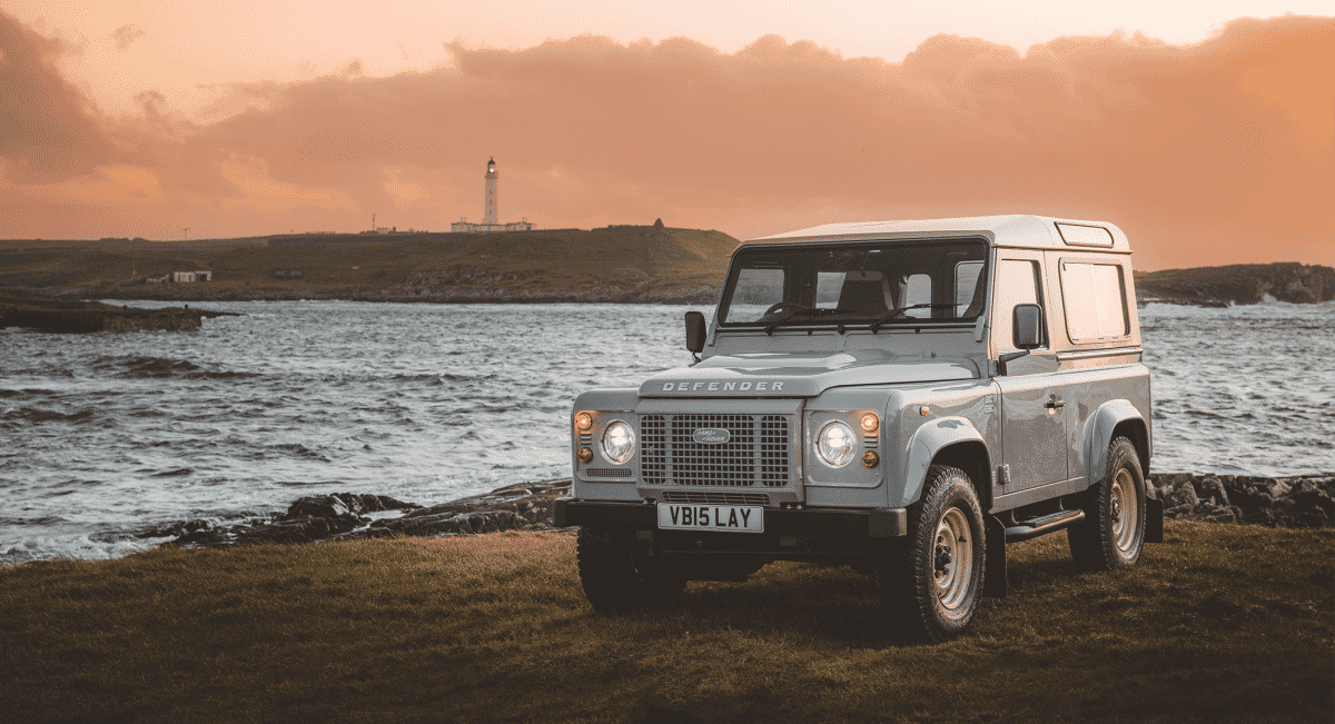 A Land Rover Defender parked on a cliffside overlooking the ocean at sunset with a lighthouse in the distance.
