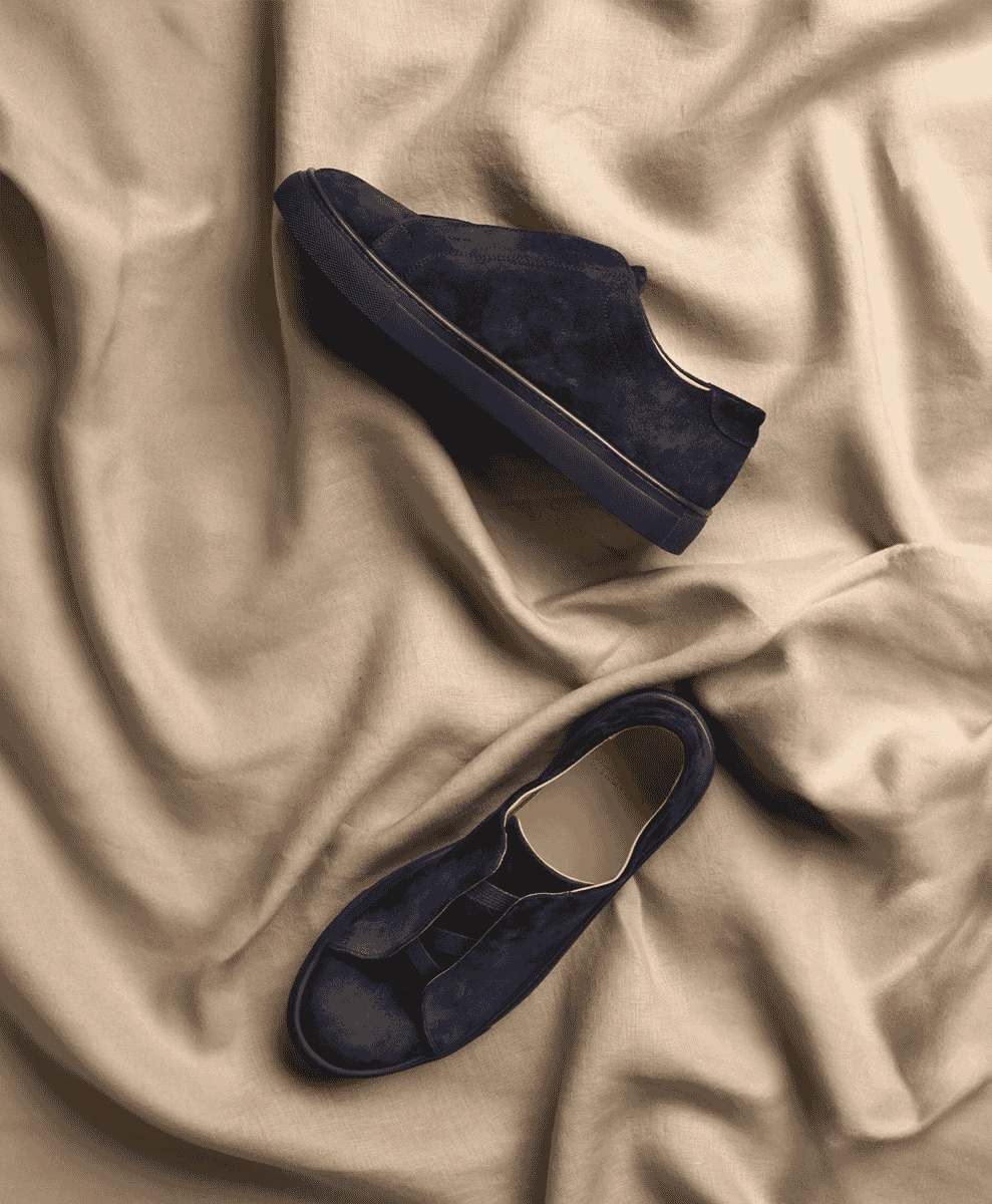Navy blue suede slip-on sneakers on a textured beige fabric background.