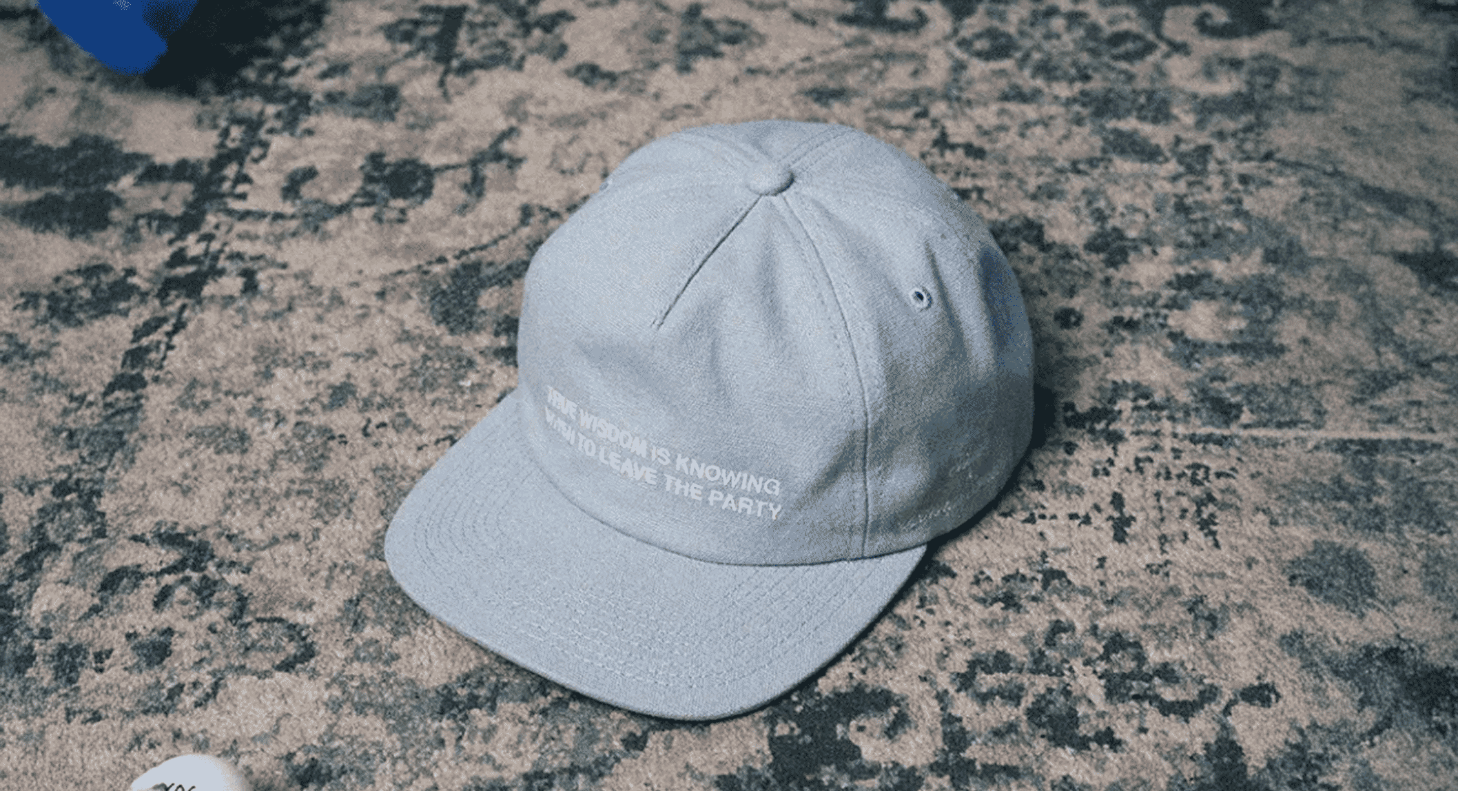 Gray Stiksen cap with 'TRUE WISDOM' text on a textured carpet.