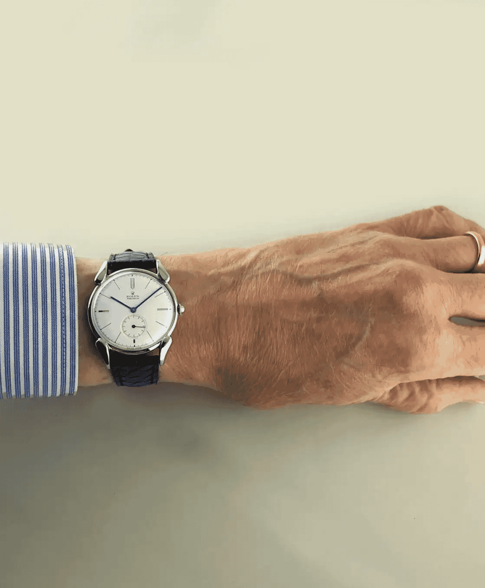 A man's wrist wearing a Rolex Precision with a white dial and black leather strap, paired with a blue-and-white striped shirt.