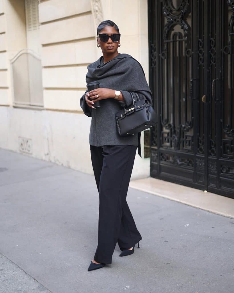 A woman in a gray top and black wide-leg trousers carrying a handbag and coffee cup on a city sidewalk.