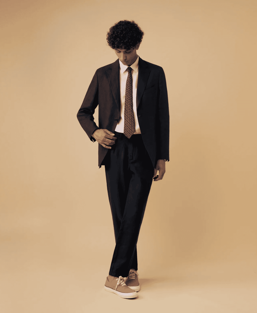 A man posing in a dark tailored suit and beige sneakers against a neutral background.