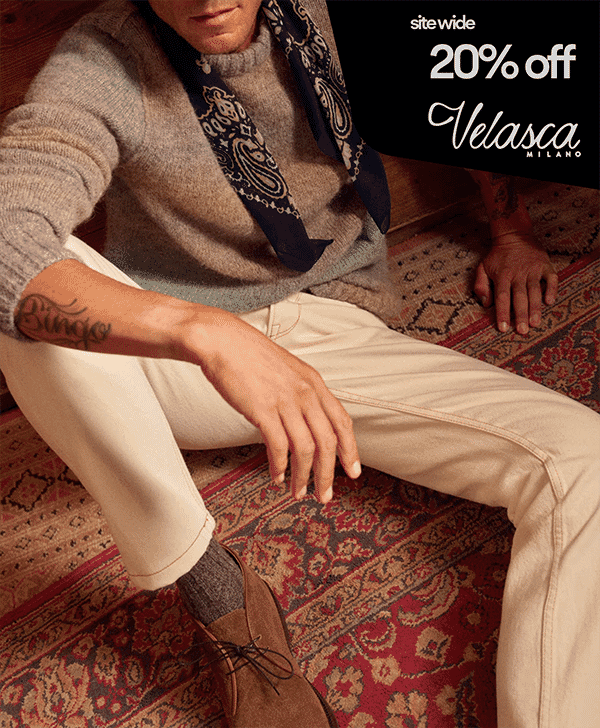 A man in a grey sweater and brown suede shoes sitting on a patterned rug.