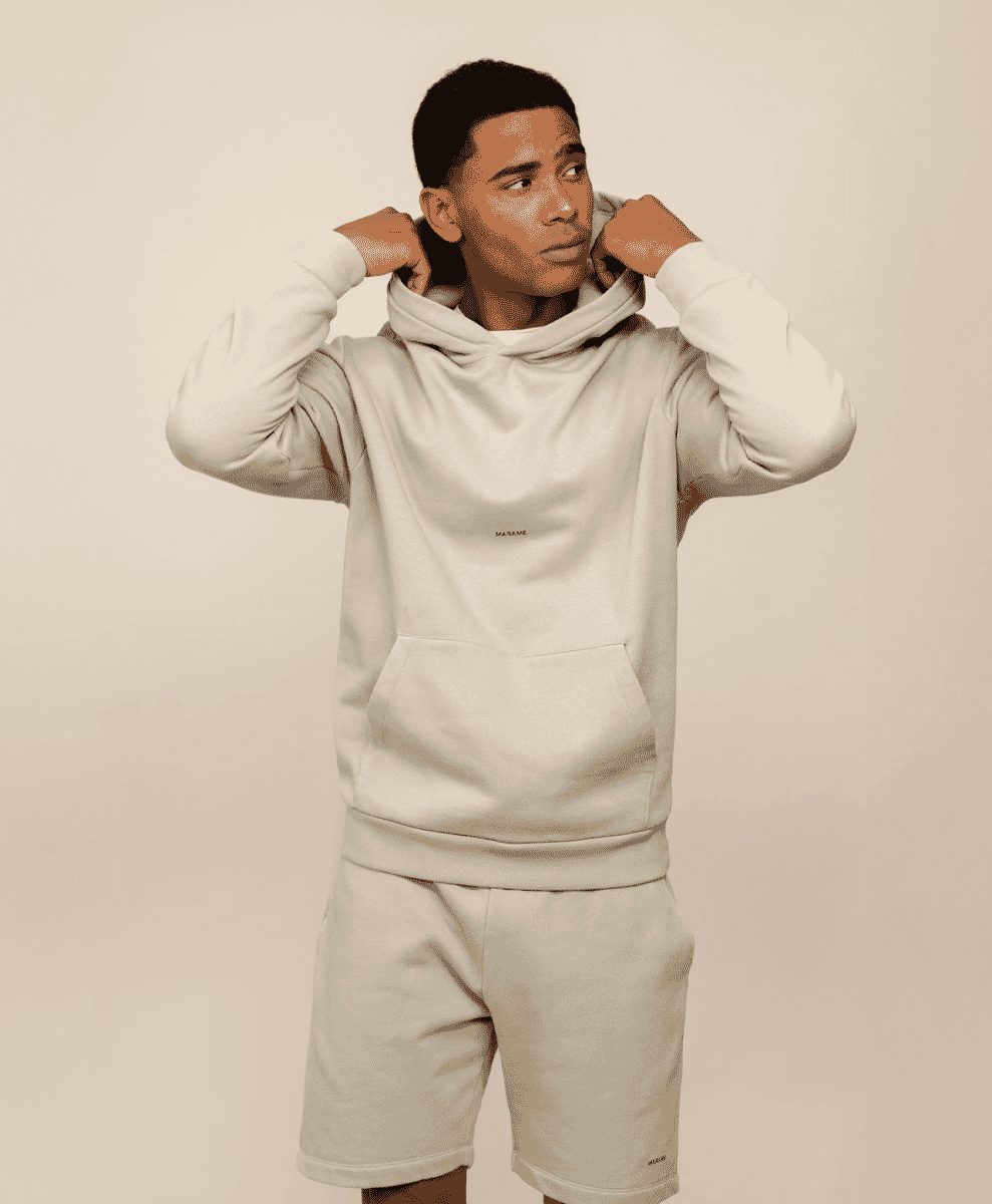 A man modeling a matching beige hoodie and shorts set in a minimalist studio.