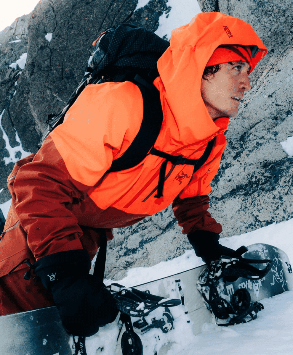 Snowboarder in a vibrant orange Arc'teryx hooded jacket navigating snowy mountain terrain.