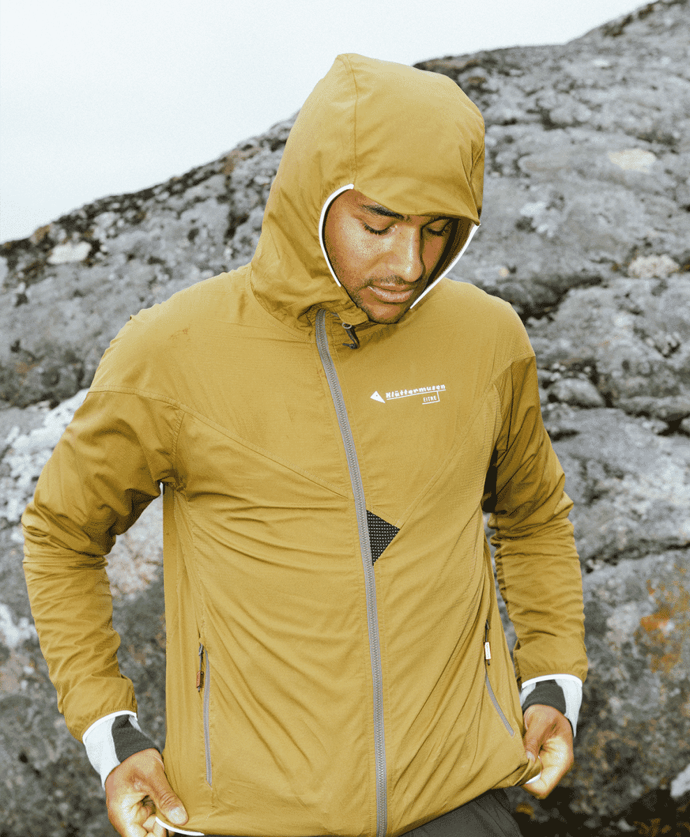 A hiker in a mustard-yellow Klättermusen technical jacket standing on a rocky mountain peak.