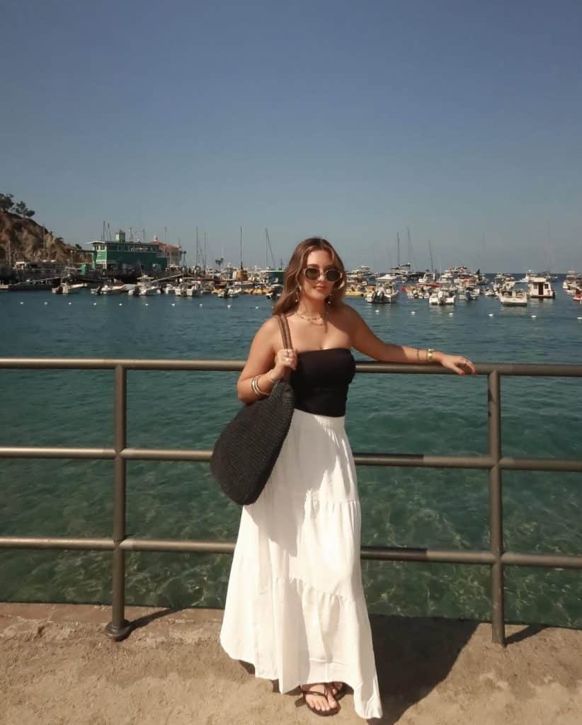 A woman in a black strapless top and white tiered maxi skirt by a harbor railing.